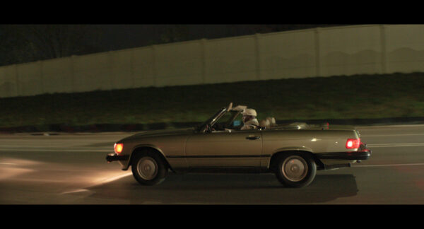A beige convertible classic car is captured cruising at night along a city roadway, its headlights illuminating the road. The driver, highlighted by passing streetlights, enjoys the open air with the car’s top down, giving the image a cinematic and nostalgic atmosphere.