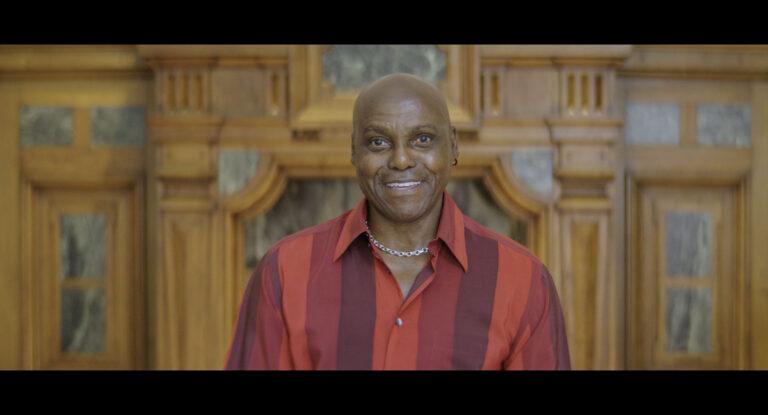 A person stands centered in front of an ornately carved wooden wall with marble inlays, wearing a bold red and burgundy striped collared shirt and a silver chain necklace. The image features warm, refined tones and a formal setting, emphasizing a dignified and polished presence.