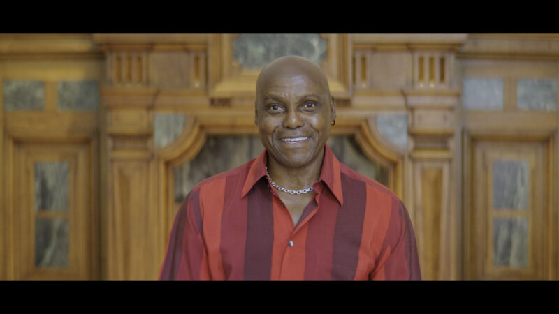 A person stands centered in front of an ornately carved wooden wall with marble inlays, wearing a bold red and burgundy striped collared shirt and a silver chain necklace. The image features warm, refined tones and a formal setting, emphasizing a dignified and polished presence.