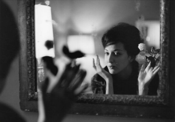 This black-and-white photograph captures a person reflected in a mirror, set in an intimate, warmly lit interior. The person is holding a white rose and lifting their hands with expressive, delicate gestures, surrounded by the soft blur of flowers and household objects in the foreground. The scene conveys a sense of introspection, artistry, and emotional nuance.
