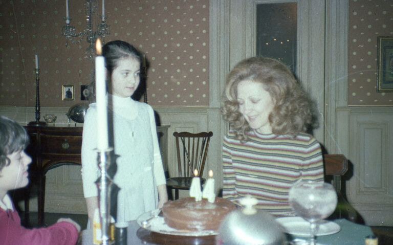 This vintage photograph captures a birthday celebration with three children gathered around a table in an ornately-papered dining room. A chocolate cake decorated with two "4" candles sits in front, while a tall white candle and glassware are set on the table. The children are dressed in attire typical of the era—one in a striped shirt, one in a dress, and one partially visible in a red sweater. The scene evokes a warm and nostalgic family atmosphere.