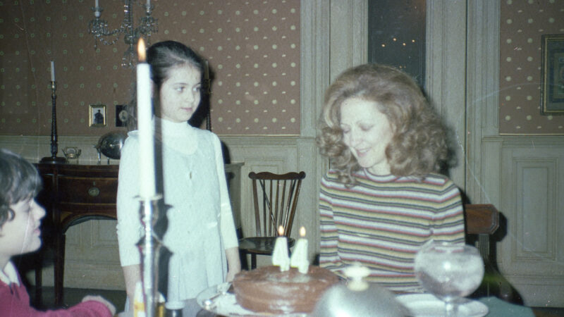 This vintage photograph captures a birthday celebration with three children gathered around a table in an ornately-papered dining room. A chocolate cake decorated with two "4" candles sits in front, while a tall white candle and glassware are set on the table. The children are dressed in attire typical of the era—one in a striped shirt, one in a dress, and one partially visible in a red sweater. The scene evokes a warm and nostalgic family atmosphere.