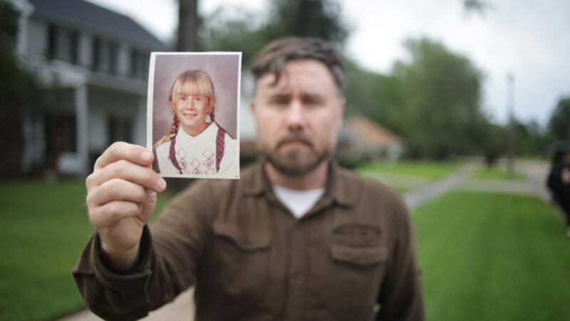 A man, facing the camera, holding a photo of a little girl.