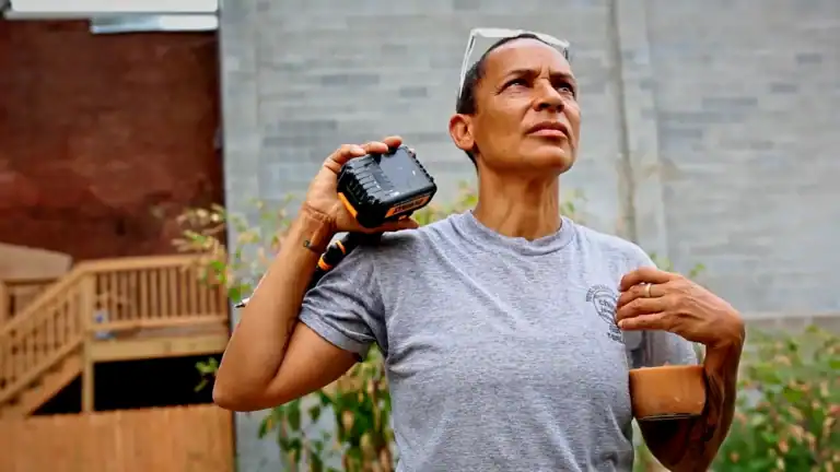 A woman holding a walkie-talkie and a coffee mug outdoors, wearing a gray T-shirt and sunglasses on her head, with a brick and concrete wall background.