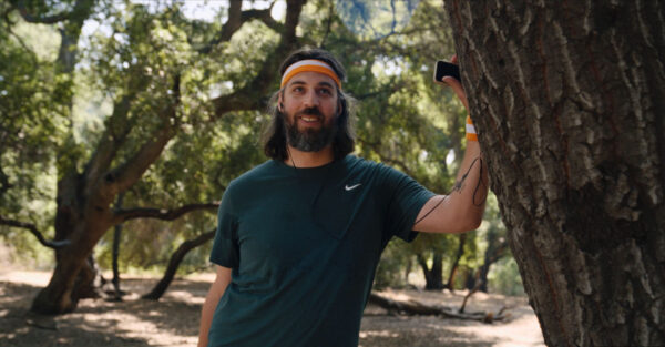 Man with long hair and beard, wearing a green Nike shirt and orange headband, leans on a tree in a forest, smiling, holding a phone.