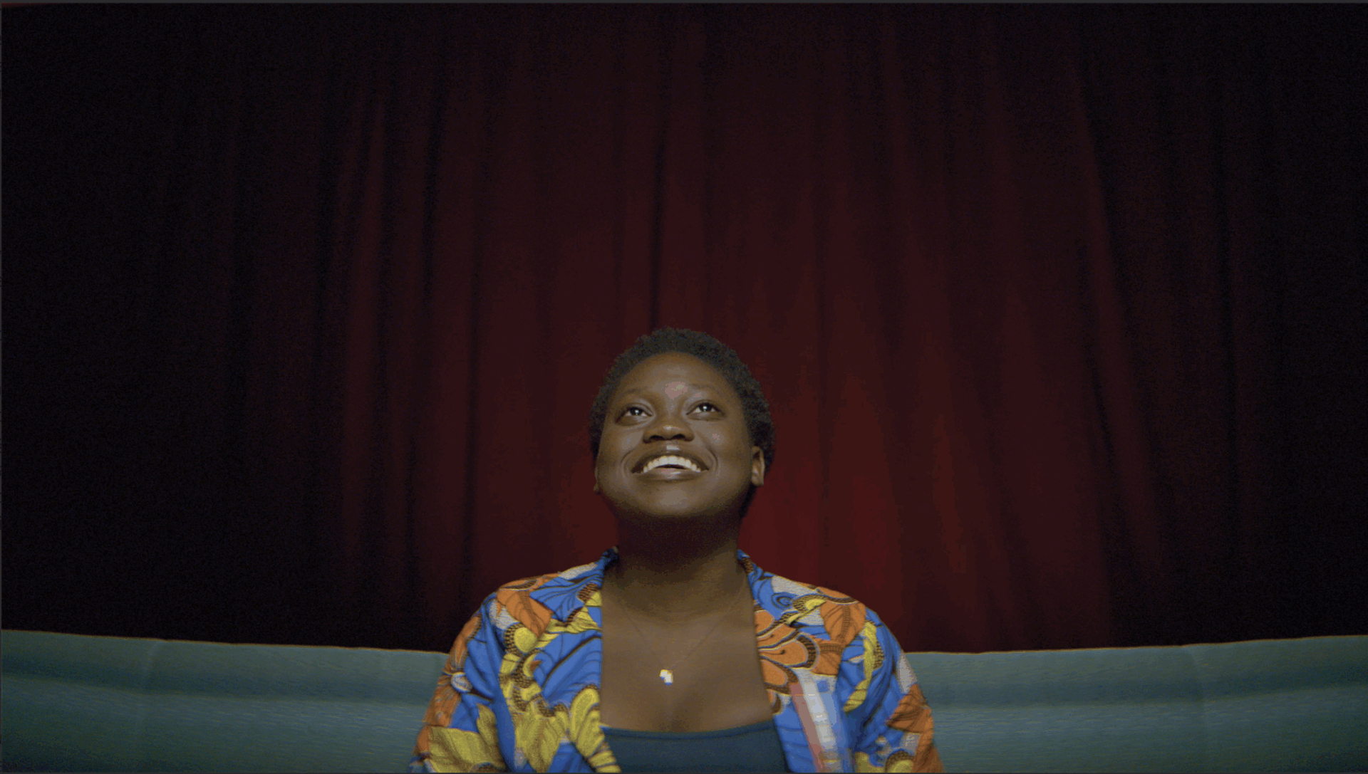 A smiling person sits in front of vibrant red curtains, wearing a colorful shirt with floral patterns, conveying a mood of joy and anticipation.