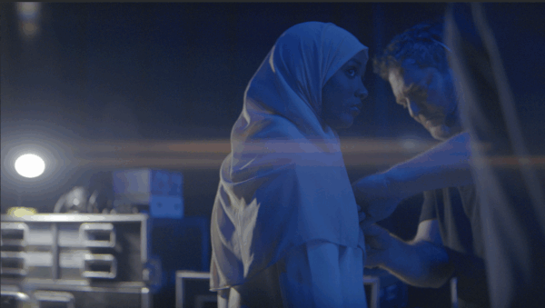 A woman in a hijab stands backstage, lit by a soft blue light. A man adjusts her clothing. The scene is calm, with a focused and supportive atmosphere.