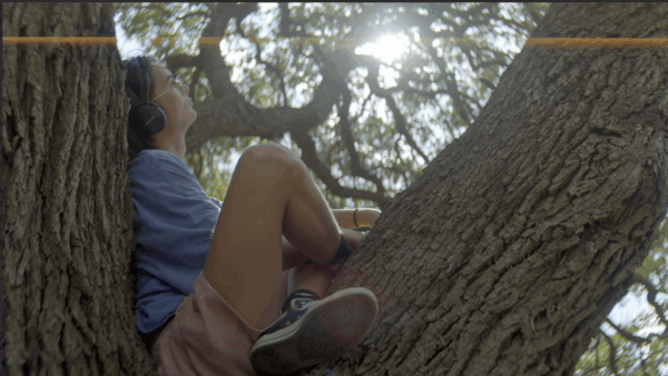 A person with headphones sits comfortably in a tree's fork, gazing up at sunlight filtering through leaves, conveying a sense of peace and relaxation.