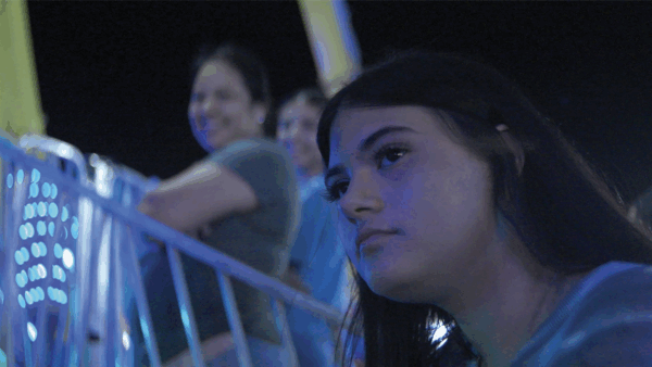 A young woman with a pensive expression rides an amusement park attraction at night, surrounded by neon lights. Behind her, people are smiling and enjoying the ride.