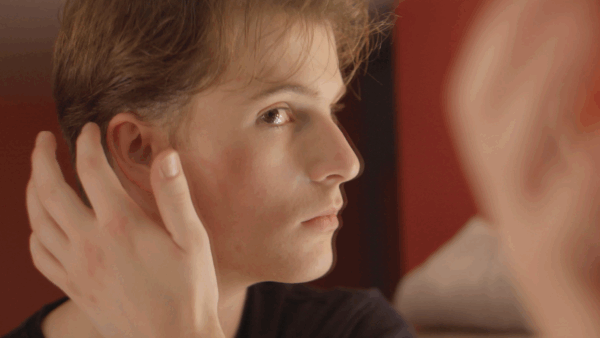 A person with short hair looks in a mirror, gently touching their head. The scene has a warm, introspective feel, with a blurred red and beige background.