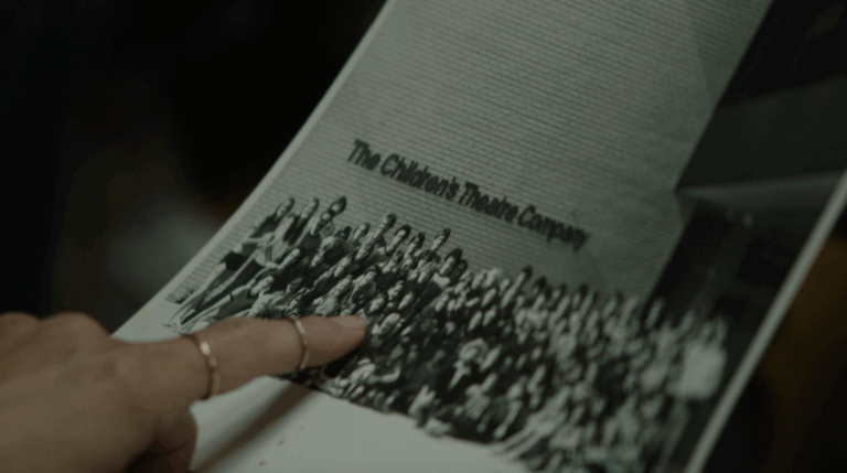 A person's hand is pointing at a group photo printed on paper. At the top of the photo, text reads "The Children's Theatre Company." The image is black and white and shows a large group of people gathered in front of a brick wall.