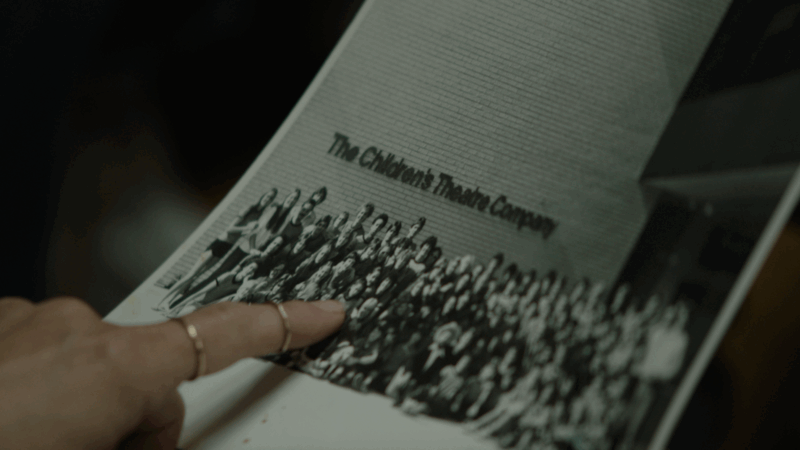 A person's hand is pointing at a group photo printed on paper. At the top of the photo, text reads "The Children's Theatre Company." The image is black and white and shows a large group of people gathered in front of a brick wall.