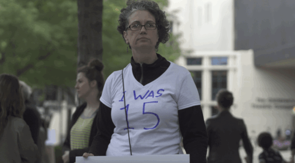 A person with short hair stands outdoors among a group of people. The person's face is blurred. They are wearing a white t-shirt with the handwritten message "I WAS 15" on the front in blue letters. The background shows trees and a building.A person with short curly hair, whose face is blurred, stands outdoors in a group of people. They are wearing a white t-shirt with blue handwritten text reading "I WAS 15." The surroundings include several people, trees, and a building in the background.