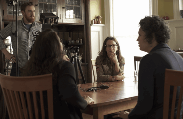 Four people are gathered around a wooden table in a warmly lit room. Three are seated with attentive expressions, while one stands beside a camera setup.