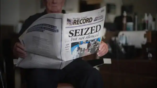 An older man sits in a barber chair reading a newspaper with the headline “SEIZED ... but not silenced.” The paper covers most of his face. The background shows barber tools and equipment.