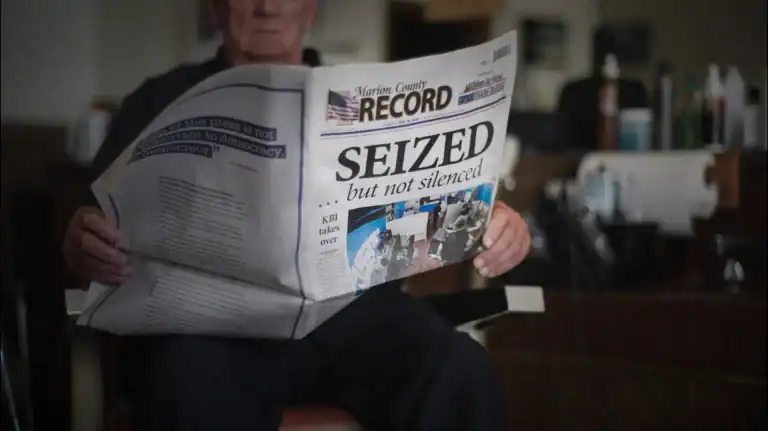 An older man sits in a barber chair reading a newspaper with the headline “SEIZED ... but not silenced.” The paper covers most of his face. The background shows barber tools and equipment.
