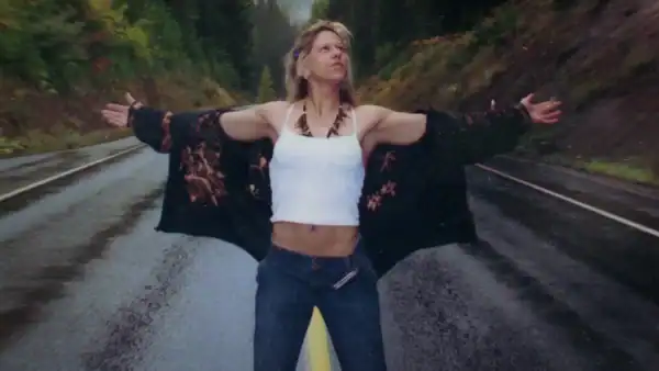 Woman standing with arms outstretched on a wet road surrounded by lush green trees, wearing a white crop top, dark jeans, and a black floral jacket, enjoying nature and freedom.