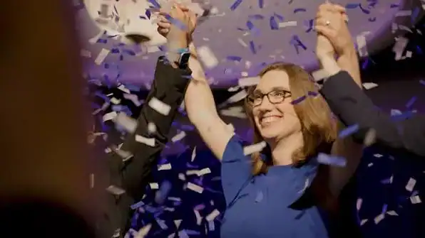 A woman surrounded by purple confetti lifts her arms in joy while holding someones hand.