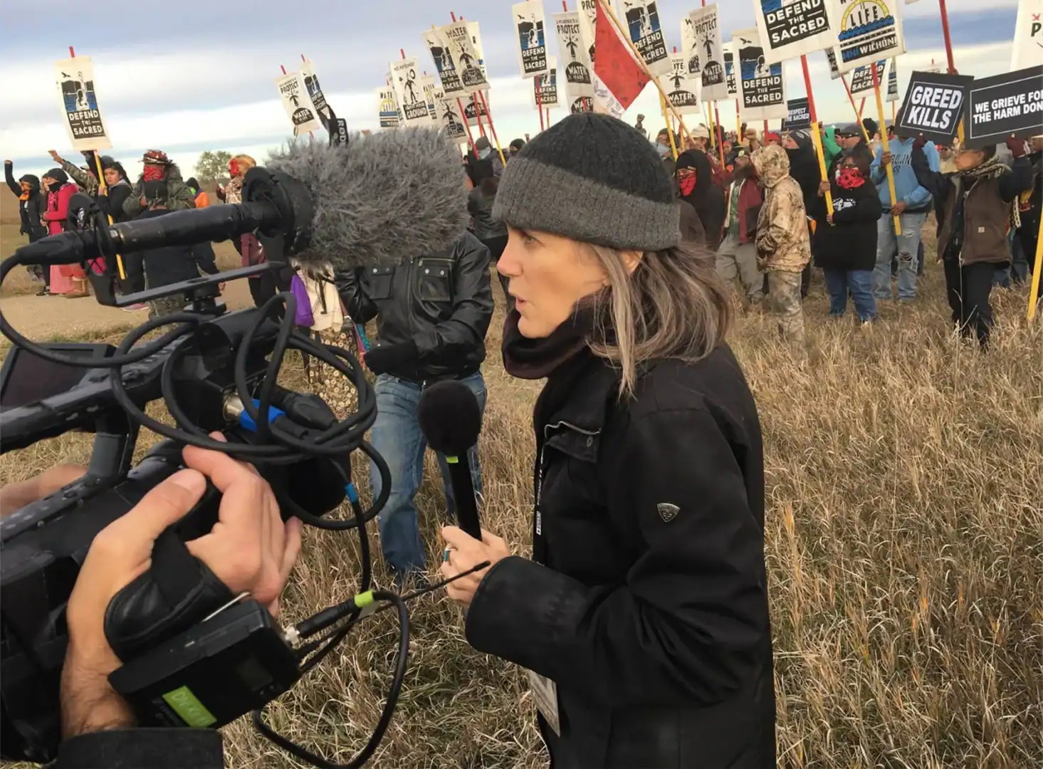 A woman holding a microphone stands in a field, surrounded by a camera crew, giving a report.