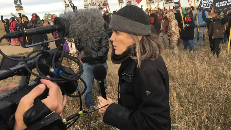 A woman holding a microphone stands in a field, surrounded by a camera crew, giving a report.