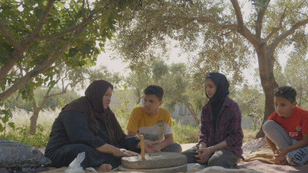 Four people sit on blankets beneath leafy trees outdoors, gathered around a traditional stone mill. One person demonstrates grinding, while the others watch or participate in conversation. Sunlight filters through the branches, and the setting feels peaceful and connected to nature.