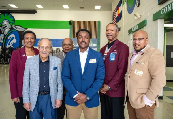 Six Tuskeegee Airmen stand together indoors, dressed in business or semi-formal attire, posing for a group photo in a brightly lit room with green, blue, and white wall accents.