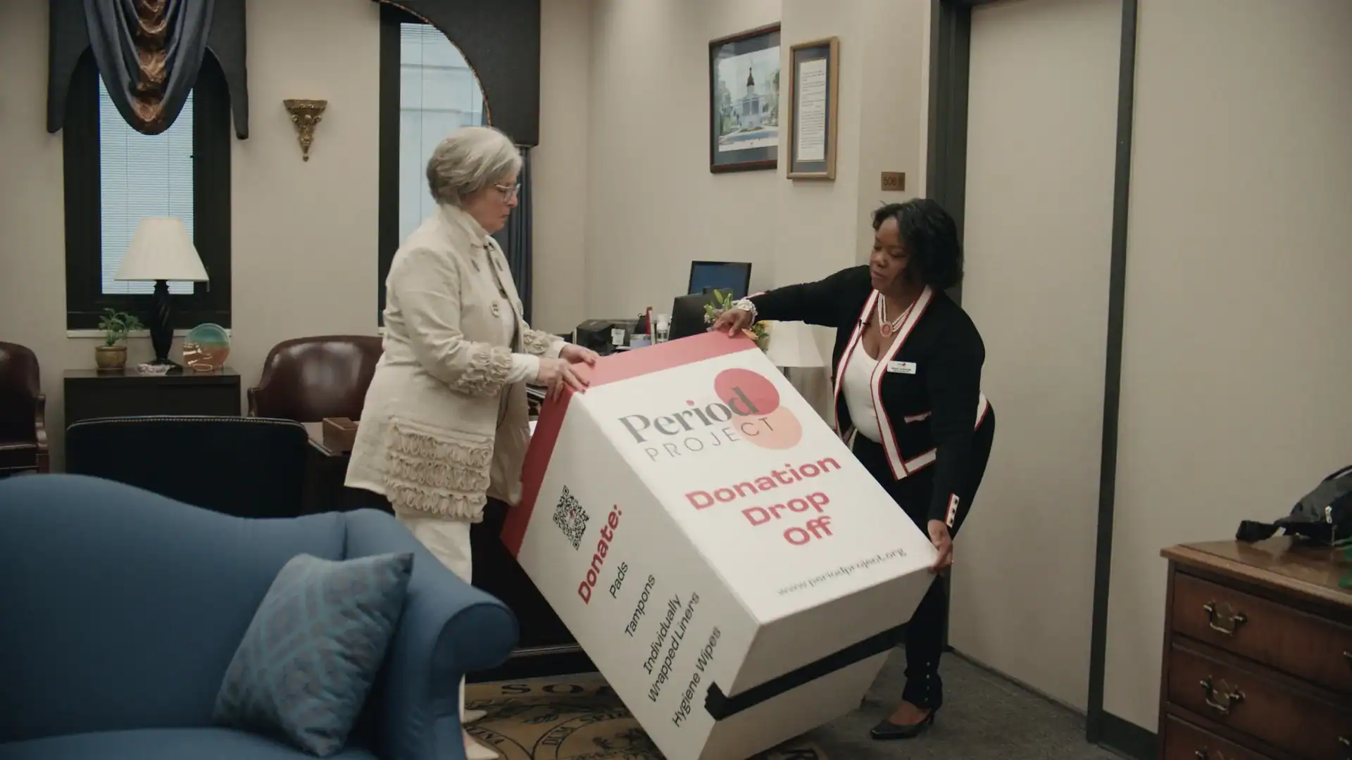 Two older women put up a donation box with the words 'Period Project' on it in an indoor setting.