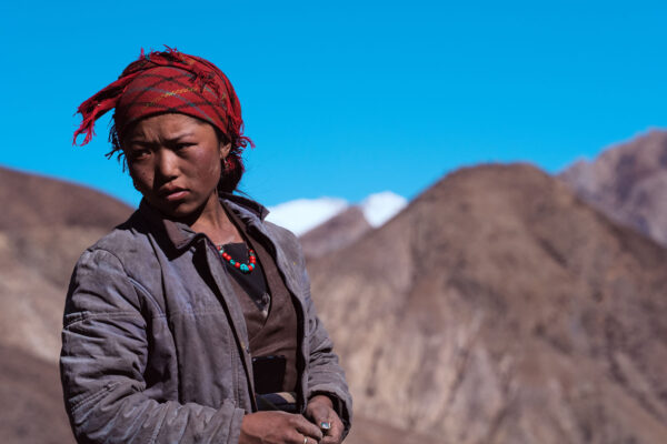 A person stands outdoors in front of brown, rocky mountains under a bright blue sky. They are wearing a red patterned headscarf, a thick gray jacket, a brown inner garment, and a necklace with colorful beads. Snow-capped peaks are visible in the distance.