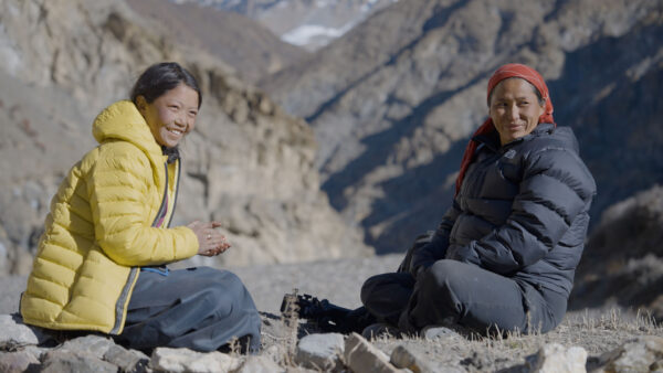 Two women sit outdoors on rocky ground in a mountain valley, dressed warmly in puffy jackets. The woman on the left wears a bright yellow coat and smiles warmly, while the woman on the right, in a dark jacket and red headscarf, looks toward her with a gentle expression. Behind them rise steep, rugged mountains under a clear sky.