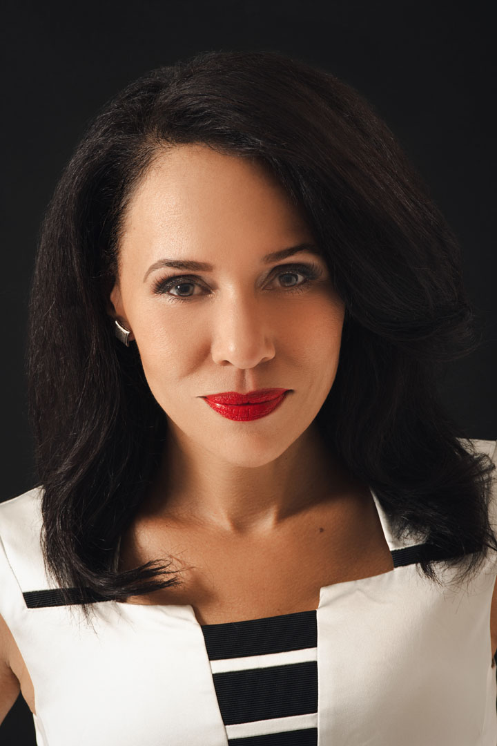 This professional portrait features a woman with shoulder-length straight dark hair wearing an elegant white dress with black accents, photographed against a plain, dark background. The lighting highlights her smooth complexion and the stylish details of her distinctive outfit.