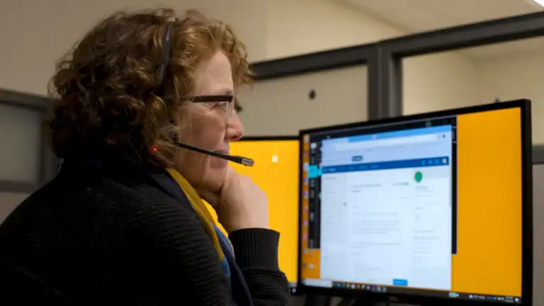 An older woman sits at a computer with a headset on.