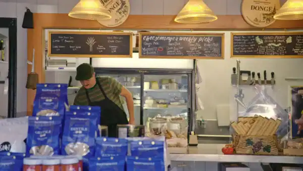 A man at a counter looks down, the background shows a fridge full of cheese.