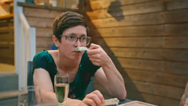 A woman with short hair wearing a green blouse, smells a cracker with cheese on it in a rustic wooden cabin setting.