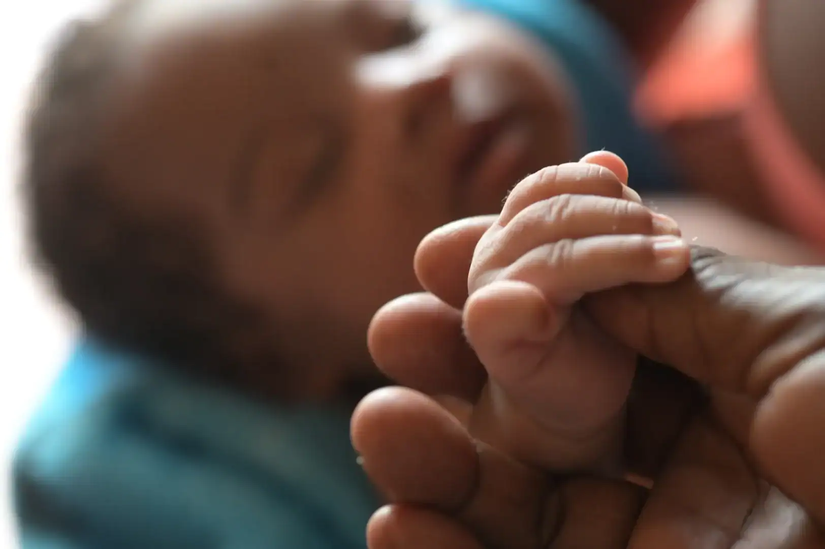 Close-up of a newborn baby's hand holding an adult's finger, symbolizing care and connection, with a blurred background of a sleeping infant.