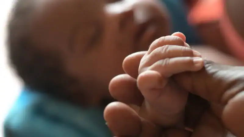Close-up of a newborn baby's hand holding an adult's finger, symbolizing care and connection, with a blurred background of a sleeping infant.