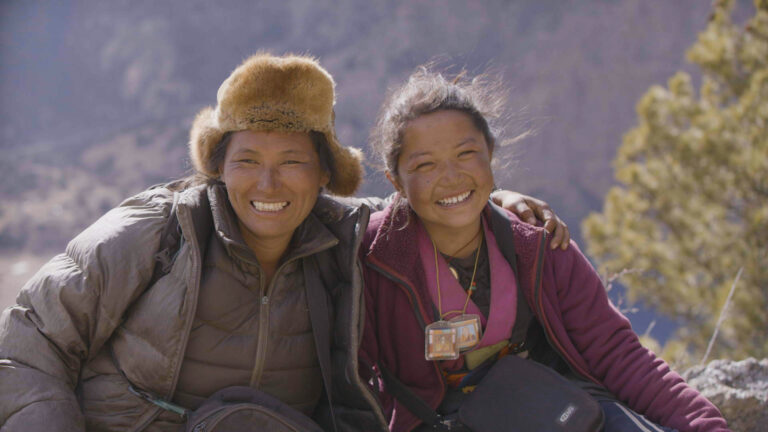 Two people sitting outdoors with mountains in the background, dressed warmly in jackets. The person on the left wears a furry hat and a grey jacket, and the person on the right wears a magenta jacket and several lanyards with photo ID badges. Both are sitting closely together, with one person's arm around the other.