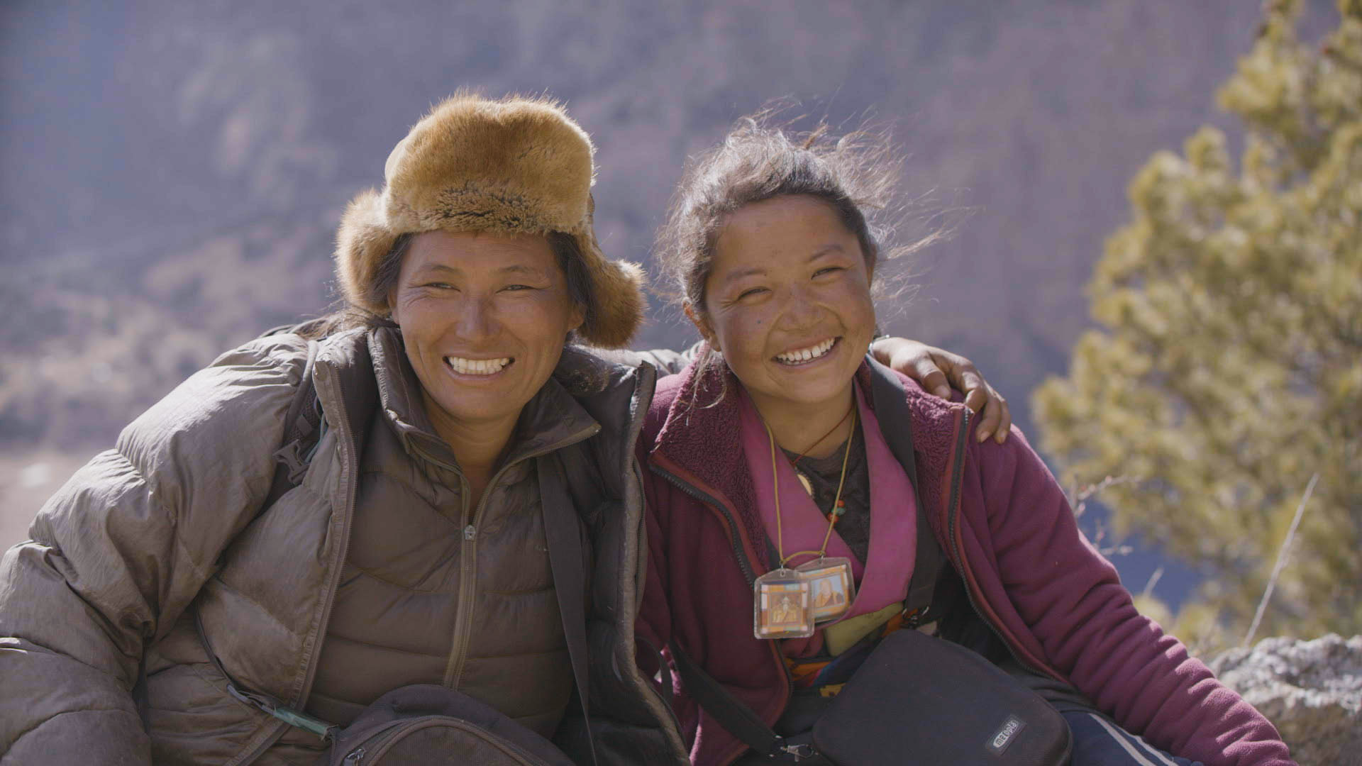 Two people sitting outdoors with mountains in the background, dressed warmly in jackets. The person on the left wears a furry hat and a grey jacket, and the person on the right wears a magenta jacket and several lanyards with photo ID badges. Both are sitting closely together, with one person's arm around the other.
