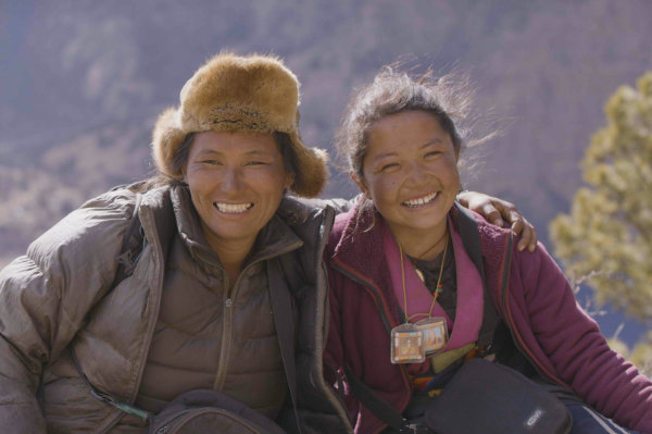 Two women wearing jackets and outdoor gear smile at the camera with mountains and trees in the background.