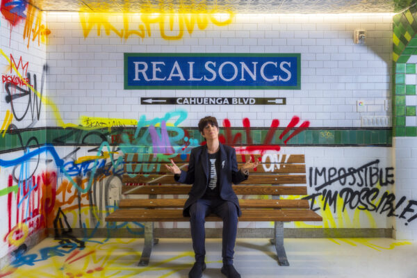 Person in dark clothing sits on a bench in a subway-themed space with a "REALSONGS" sign and colorful graffiti on the tiled walls.
