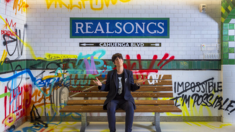 Person in dark clothing sits on a bench in a subway-themed space with a "REALSONGS" sign and colorful graffiti on the tiled walls.