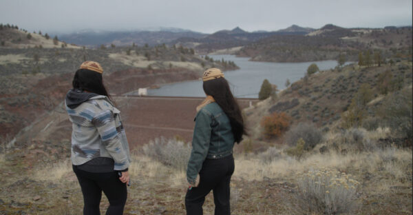 Two women stand on a hillside overlooking a reservoir and dam, surrounded by dry grass, shrubs, and rolling hills. Both wear patterned beanies and jackets, with their backs turned toward the camera, gazing at the water and distant mountains under a cloudy sky.