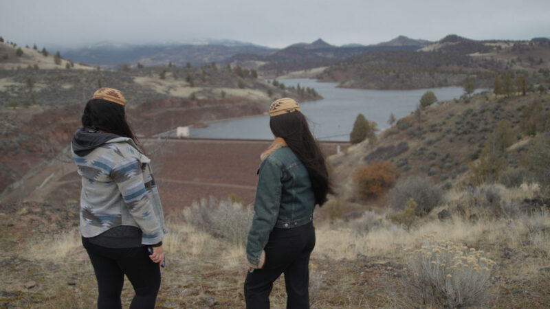 Two women stand on a hillside overlooking a reservoir and dam, surrounded by dry grass, shrubs, and rolling hills. Both wear patterned beanies and jackets, with their backs turned toward the camera, gazing at the water and distant mountains under a cloudy sky.