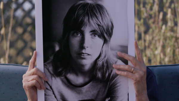 A person holds a black-and-white photo of a woman with a serious expression and shoulder-length hair.