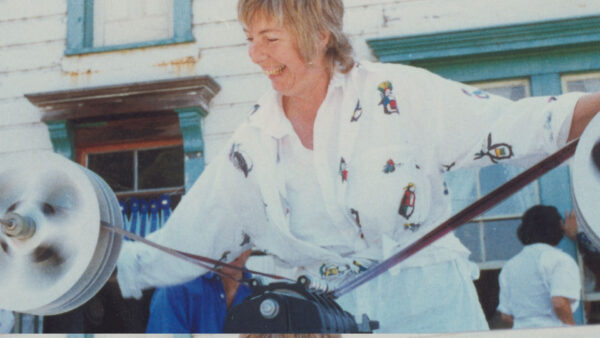 A woman joyfully spins wool on a spinning wheel outside a rustic building. She wears a patterned shirt, conveying a vibrant and lively atmosphere.