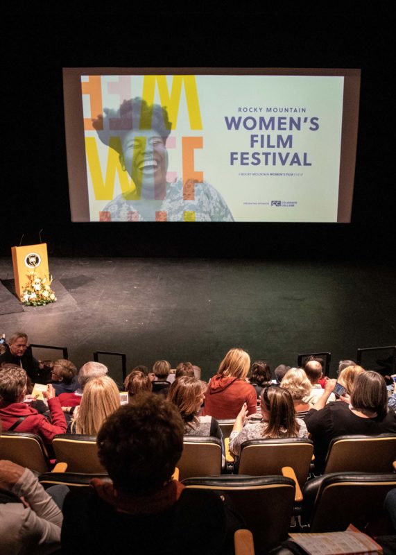A large audience seated in a darkened theater auditorium facing a stage with a podium decorated with flowers. Above the stage, a screen displays a colorful graphic for the Rocky Mountain Women's Film Festival. The crowd appears attentive and engaged, with some looking at programs while awaiting the event to begin.
