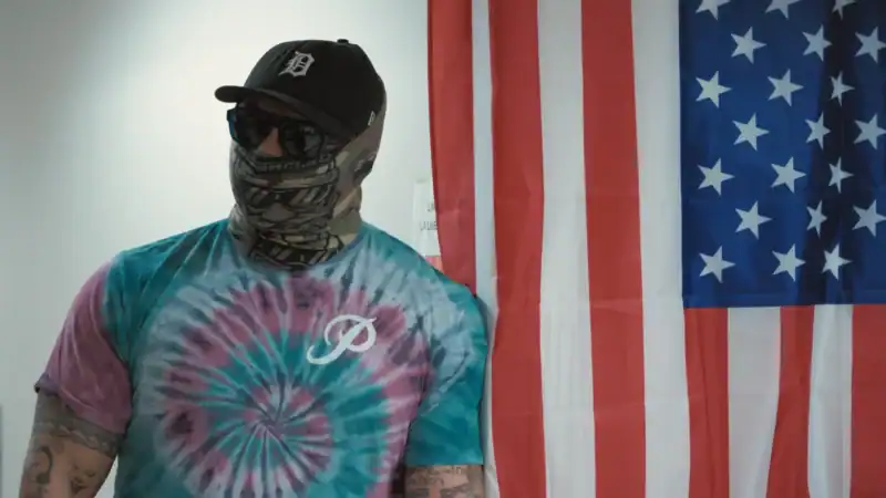 Man in a ski mask, baseball cap, and sunglasses wearing a tye-dyed shirt, looks off to his left with an American flag in the back.