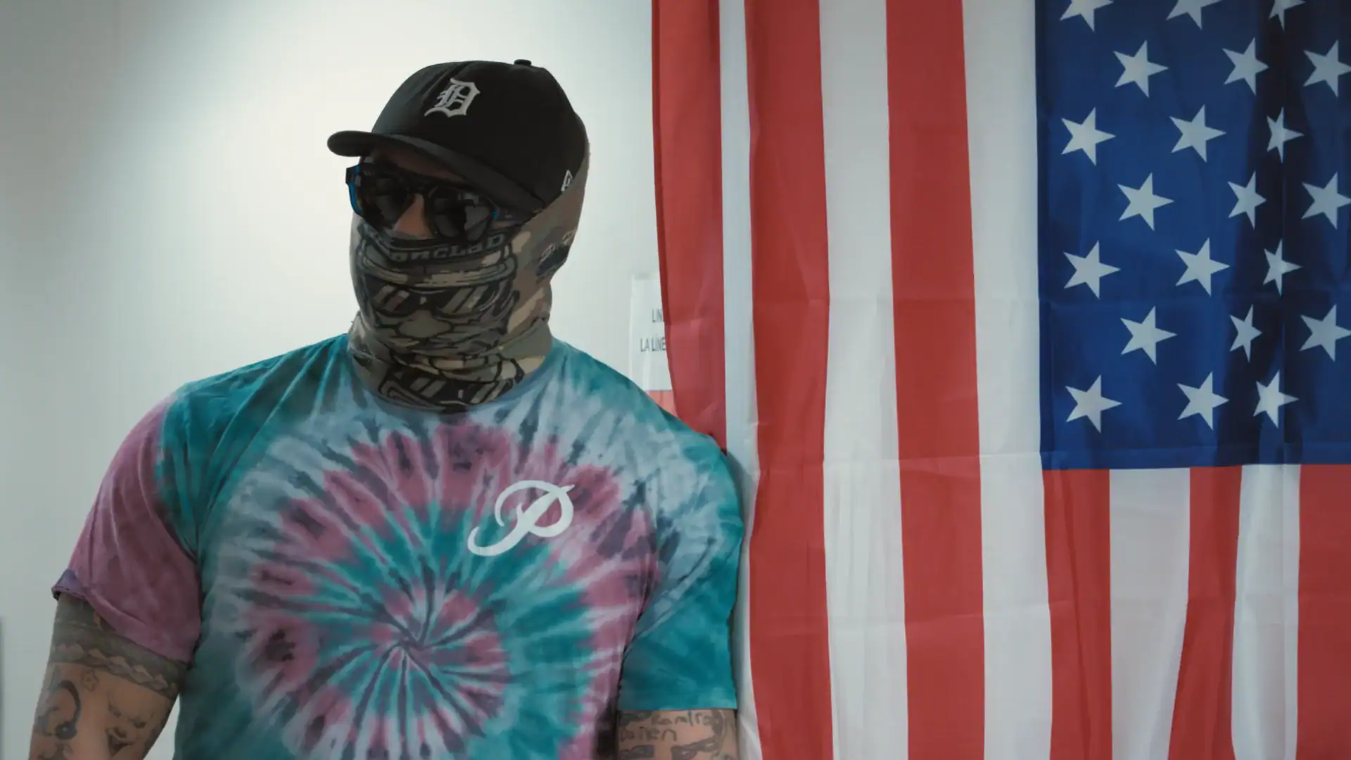 Man in a ski mask, baseball cap, and sunglasses wearing a tye-dyed shirt, looks off to his left with an American flag in the back.