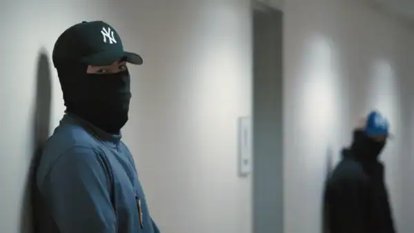 Person wearing a black ski mask and blue jacket standing against a wall, with another person in the background in a hallway setting.