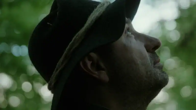 Close-up of a man wearing a hat, looking upward in a lush green outdoor setting, capturing a moment of reflection or admiration in nature.