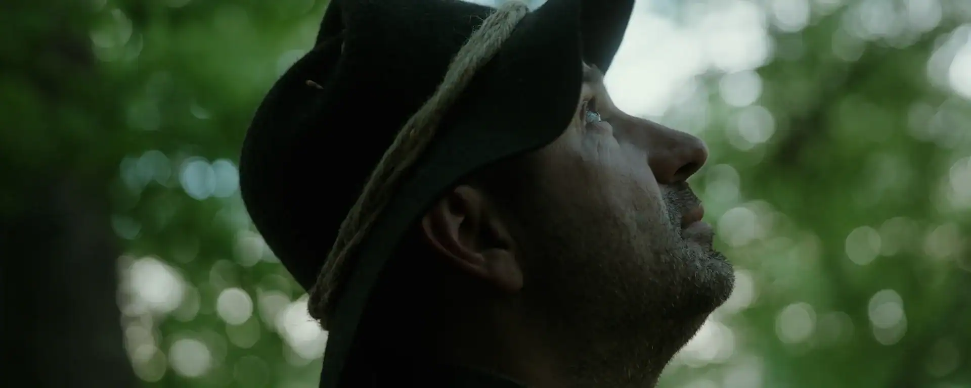 Close-up of a man wearing a hat, looking upward in a lush green outdoor setting, capturing a moment of reflection or admiration in nature.